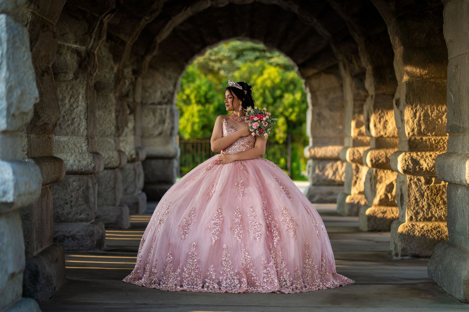 A person in a pink quinceañera ball gown poses under a stone archway with sunlit greenery in the background.