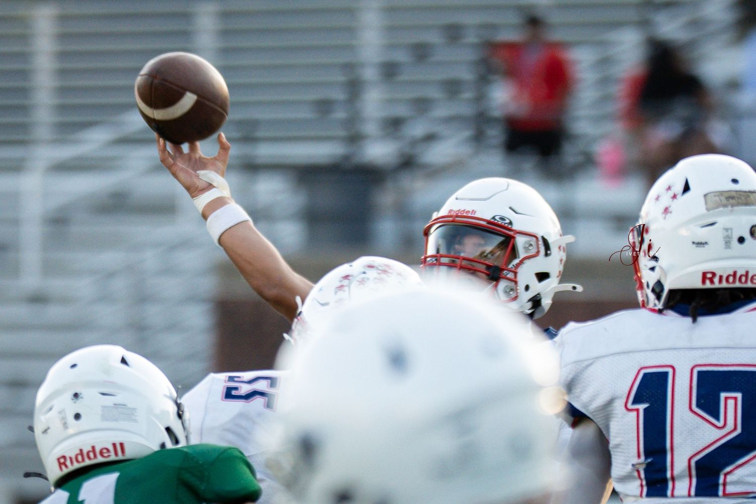 A football player in a white jersey holds the ball high above defenders during a game.