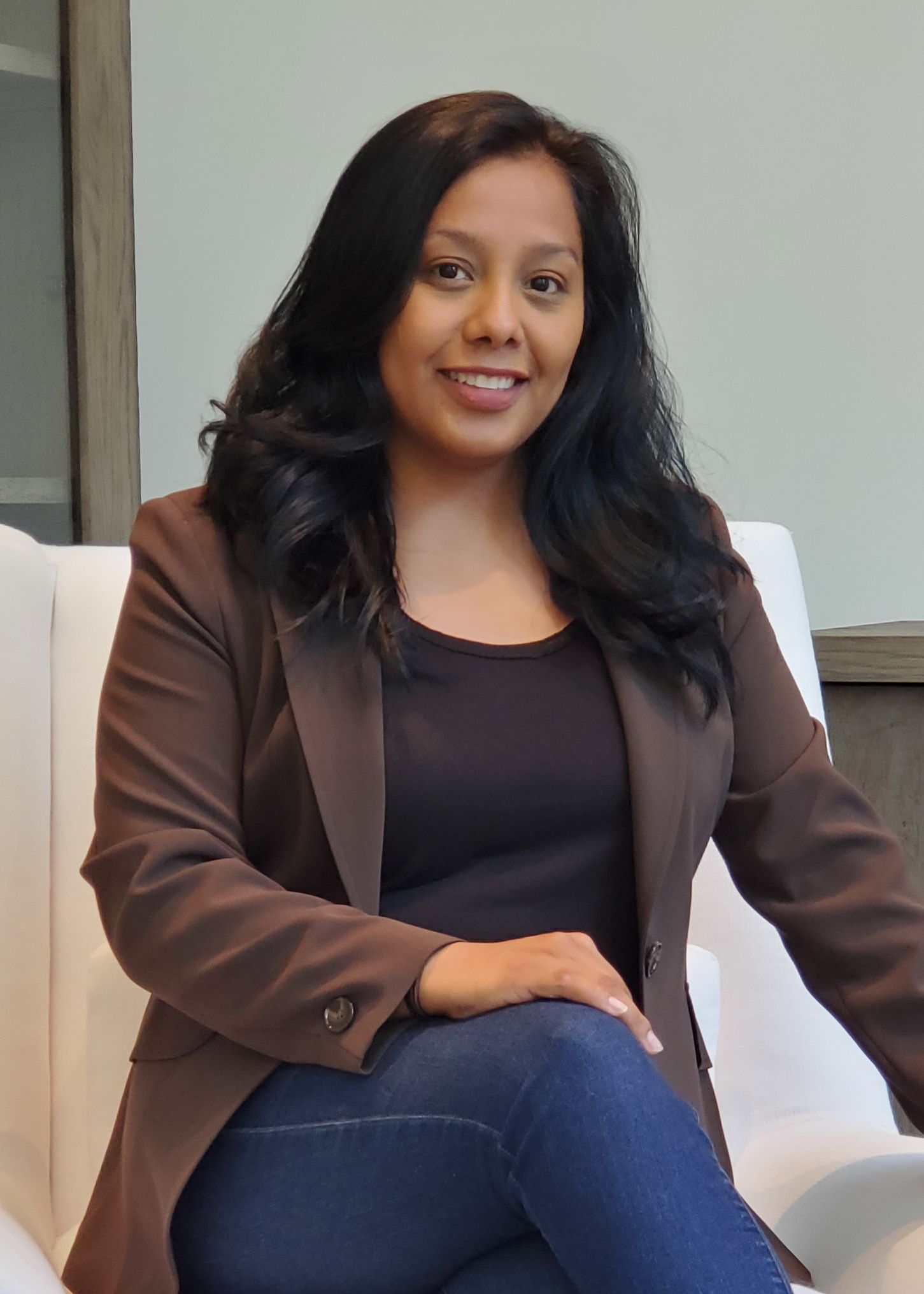Professional wearing a brown blazer and black top seated in a white chair with a polished business look.