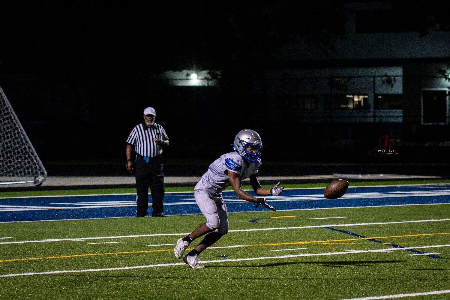 Football player in white uniform catches a pass on illuminated blue turf field at night with referee in background.