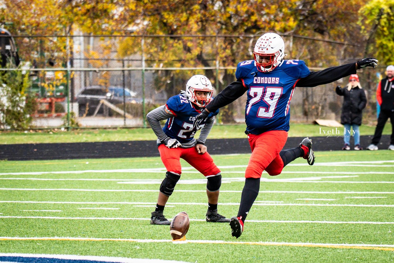 Football players from opposing teams collide during a tackle attempt on autumn afternoon with fall foliage visible.