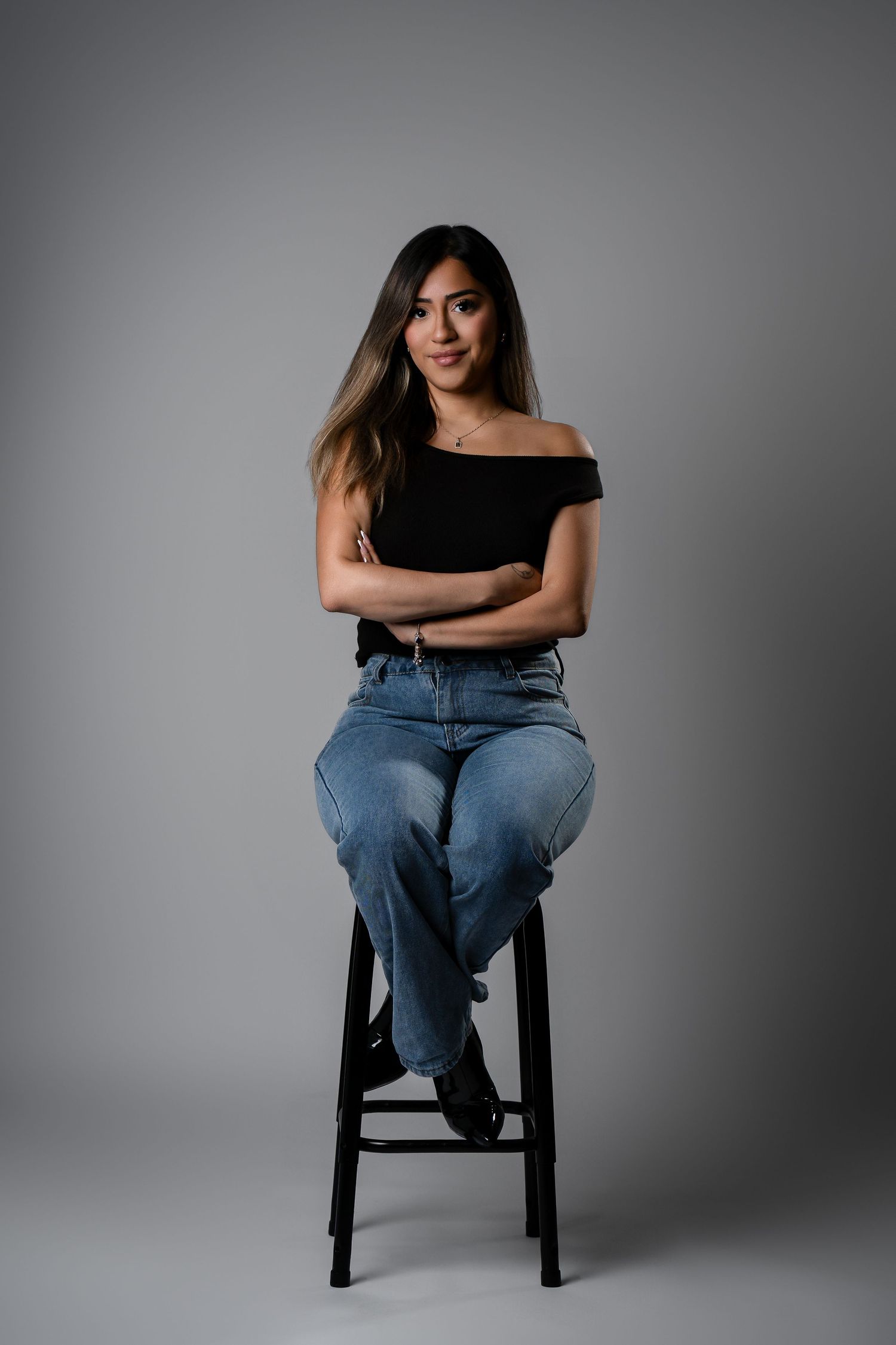 Casual studio portrait with black off-shoulder top and jeans on wooden stool against grey background.