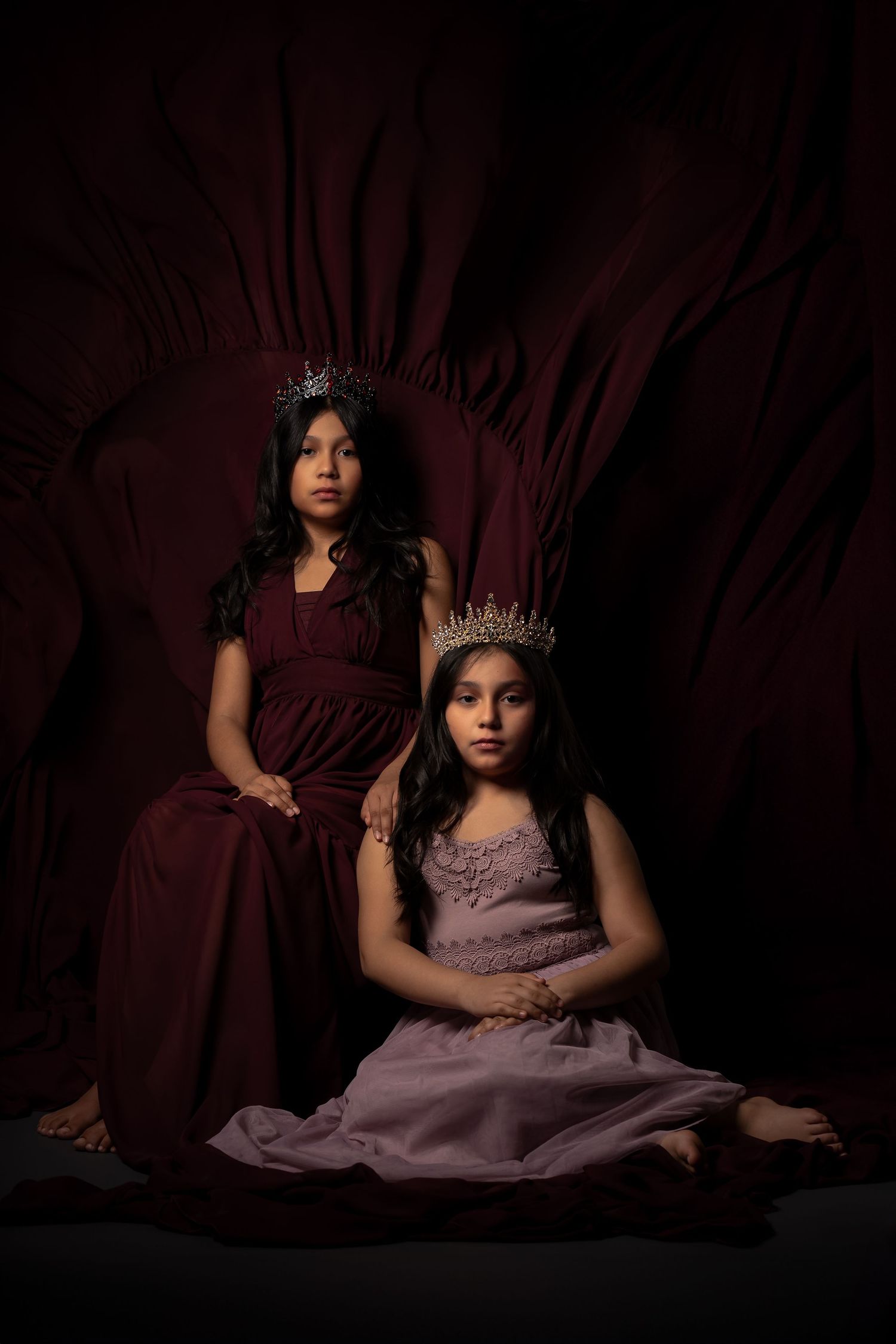 Regal portrait with crowns and formal dresses in deep burgundy and lavender against dark backdrop.