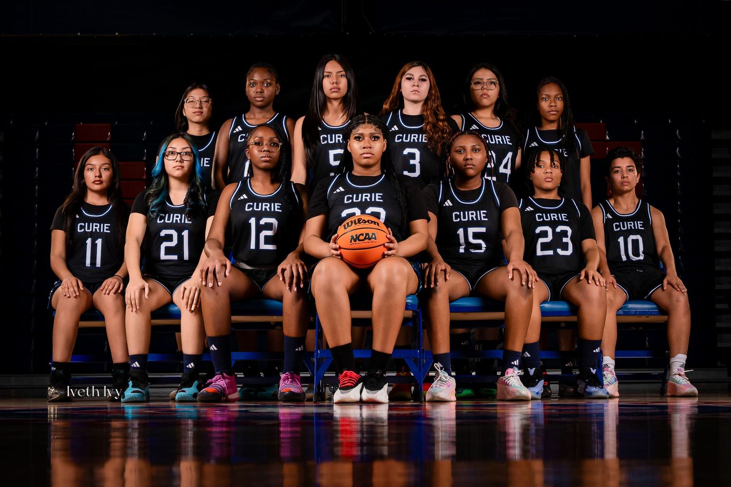 Group of basketball players in dark uniforms sitting in a row for team photo against black background.