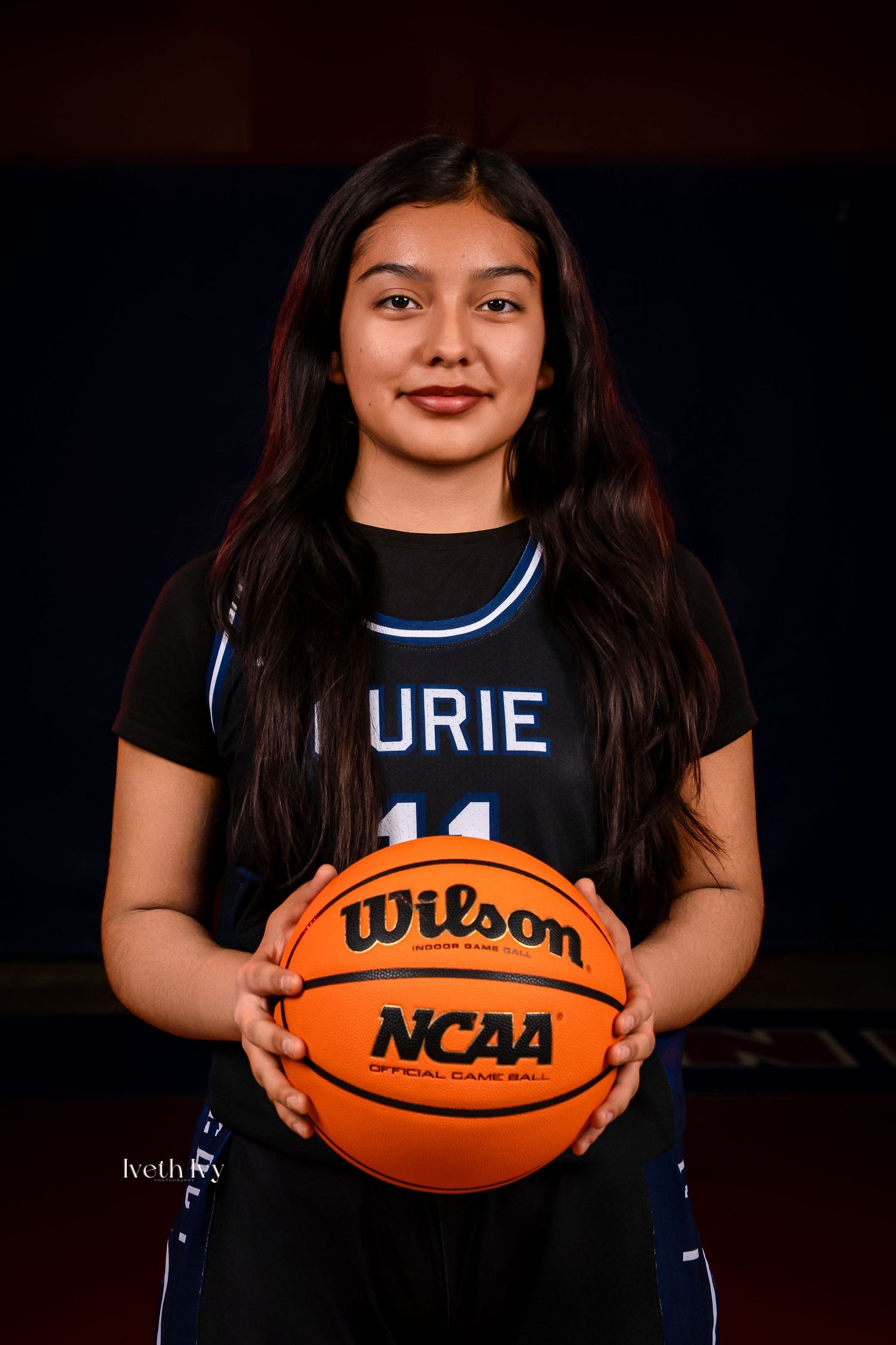 Basketball player poses with NCAA ball in dark studio setting with dramatic lighting and black uniform.