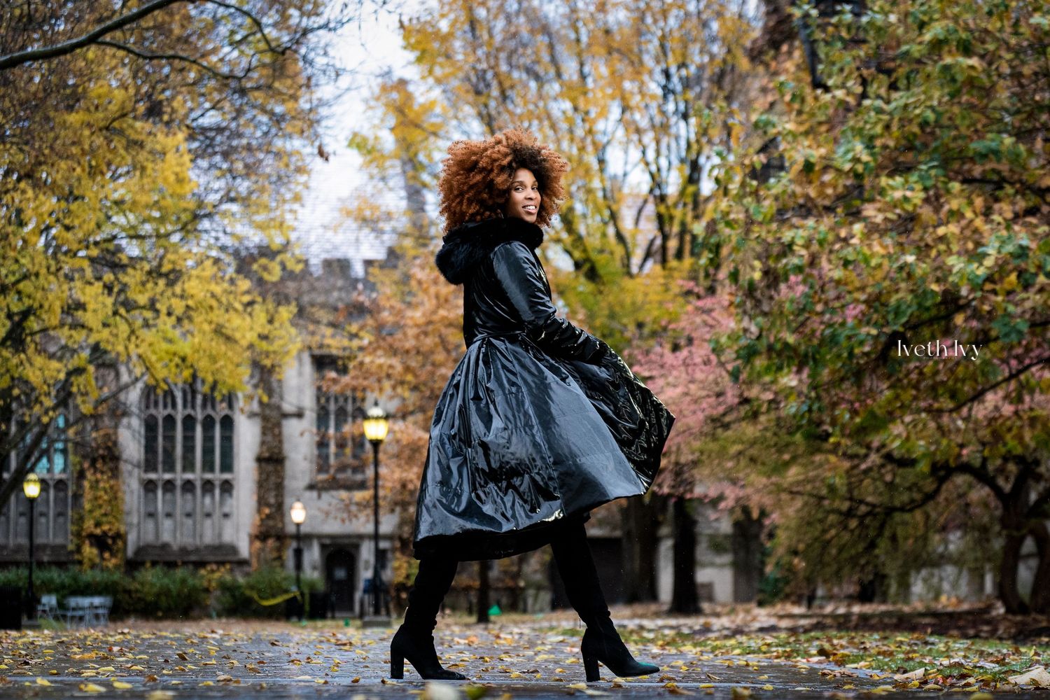 Woman in black shiny coat and boots twirling outdoors among autumn trees and fallen leaves.