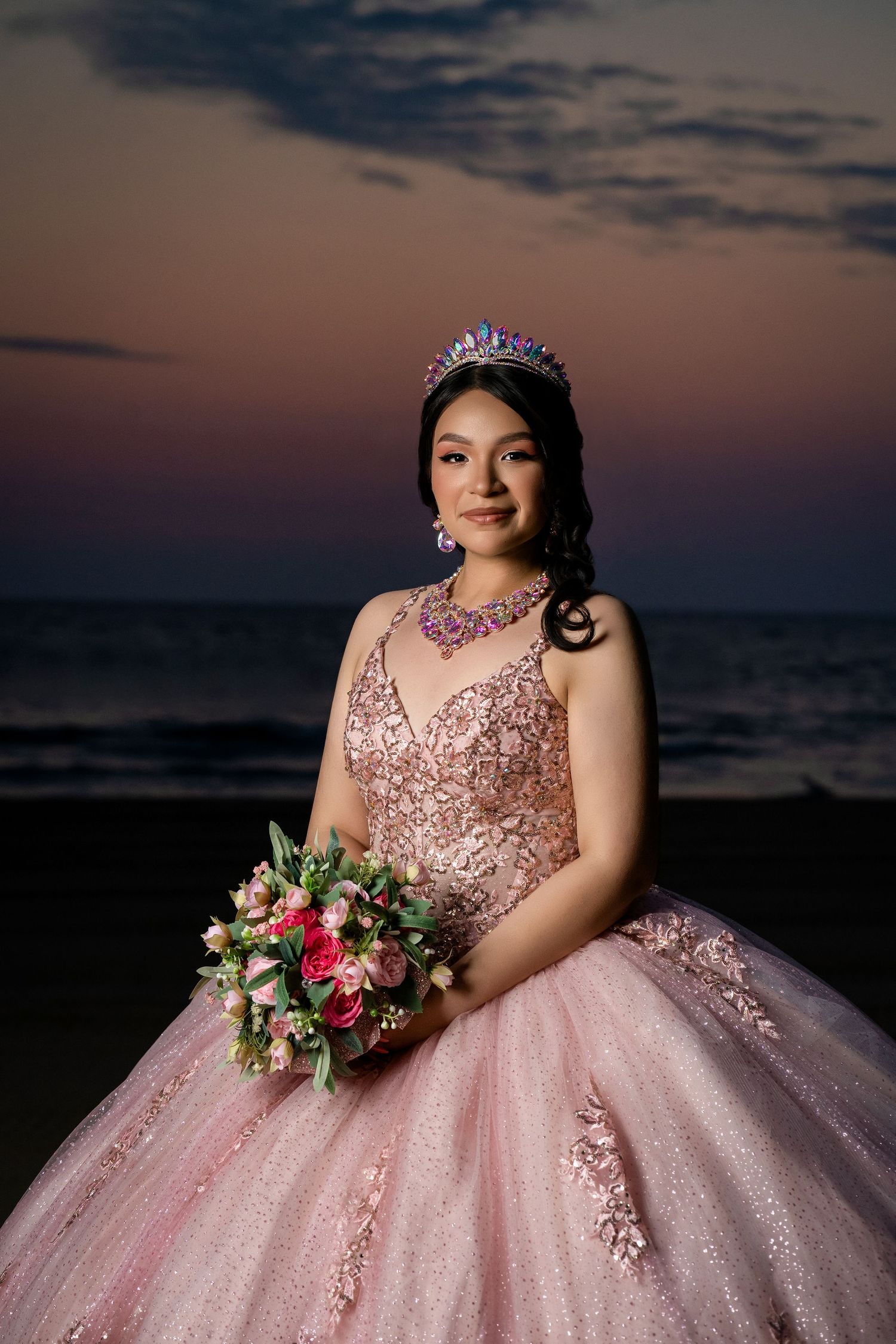 A quinceañera photoshoot at sunset featuring a sparkly pink ball gown and floral bouquet against a dusky sky.