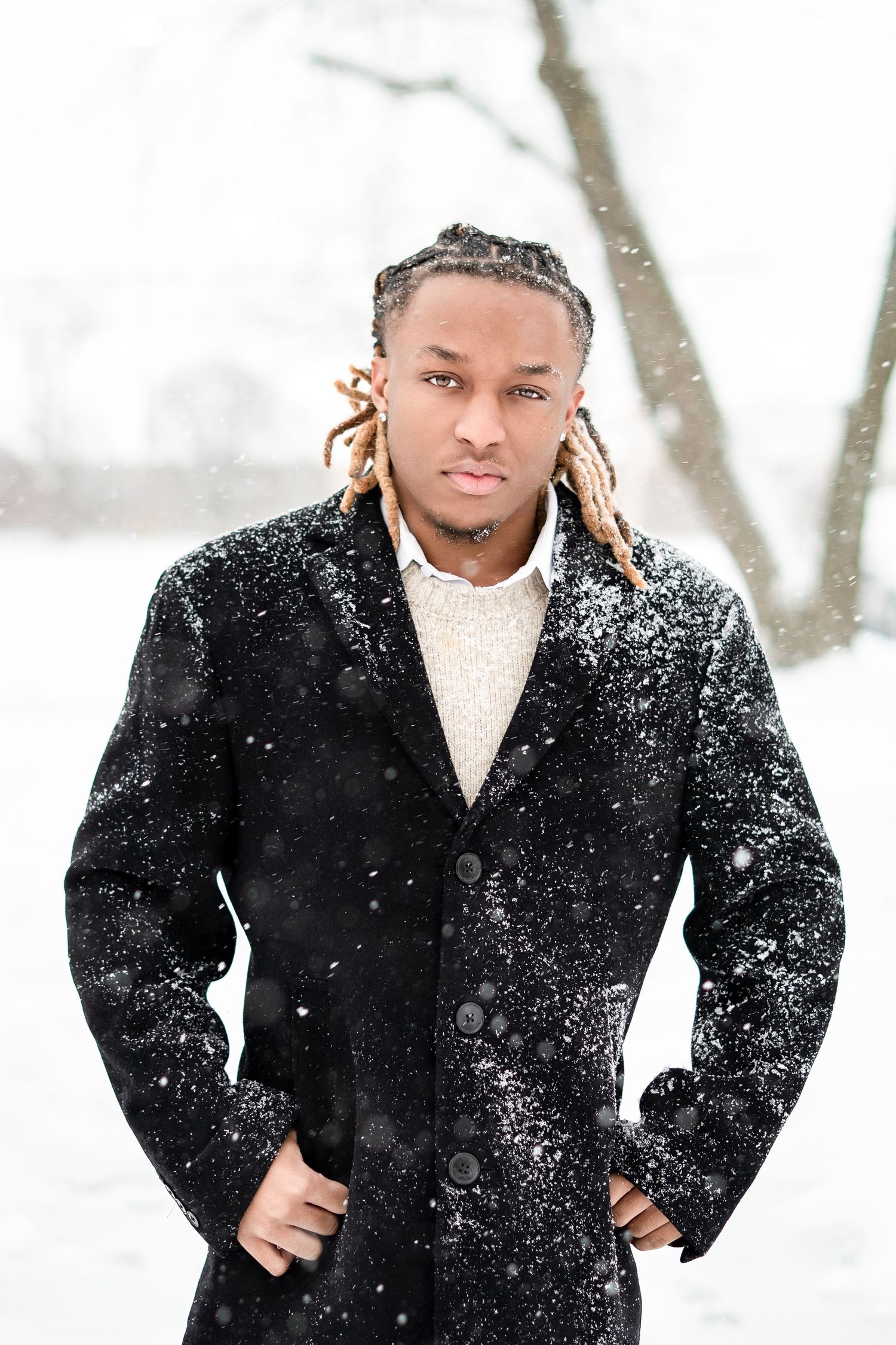 Young Black man with dreadlocks wearing a black coat standing outdoors in a snowstorm.