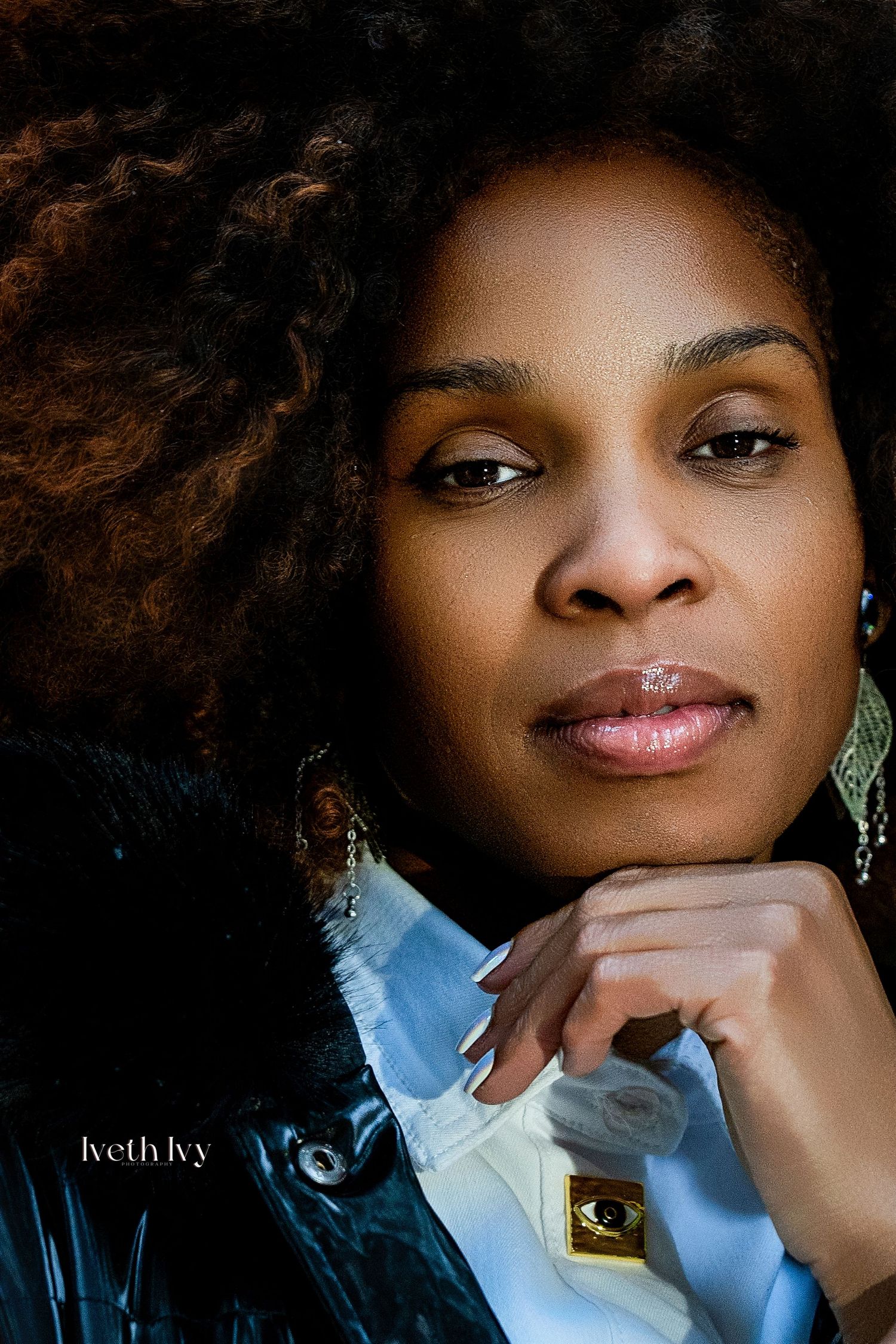 A Black woman with voluminous natural hair poses thoughtfully, wearing dangling earrings and a blue shirt, photographed by Iveth Ivy.