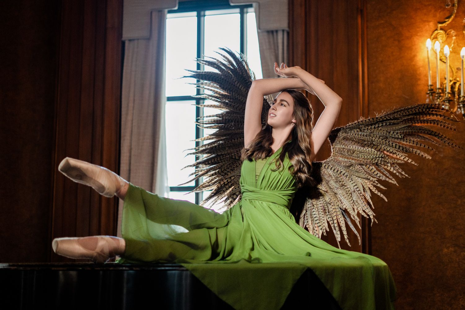 Ballet dancer in flowing green dress leaps gracefully across grand piano in ornate room with chandelier.