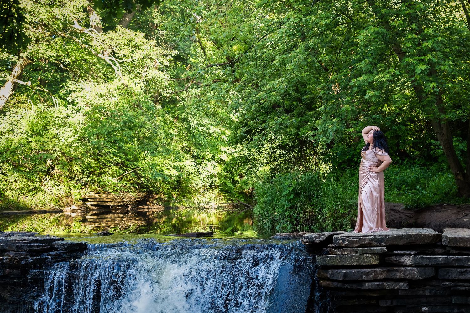 A person in a formal gown stands atop a stone wall near a cascading waterfall surrounded by lush green forest.