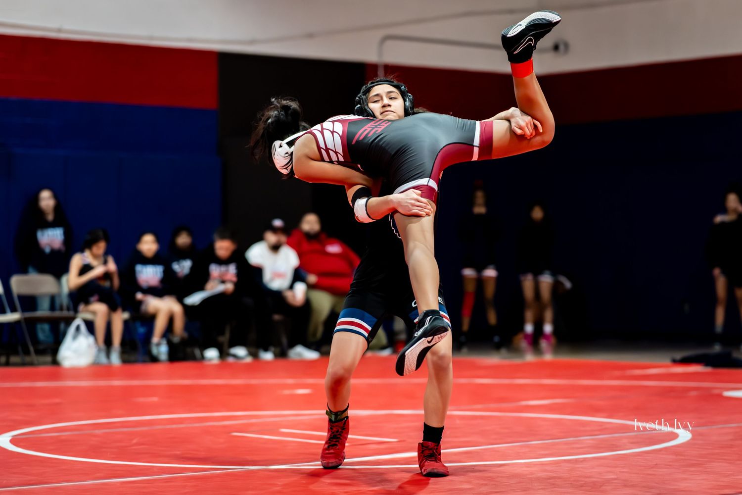 Two wrestlers engage in a competitive match on a red wrestling mat during a tournament.