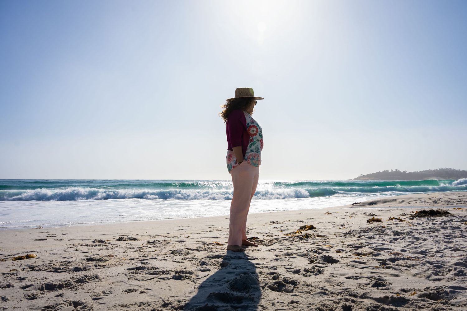 Woman in hat and shorts standing on sunny beach with turquoise ocean waves in background.