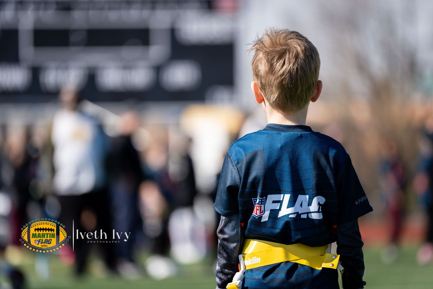 Young boy in FLAG football jersey watches players on field during youth flag football game.