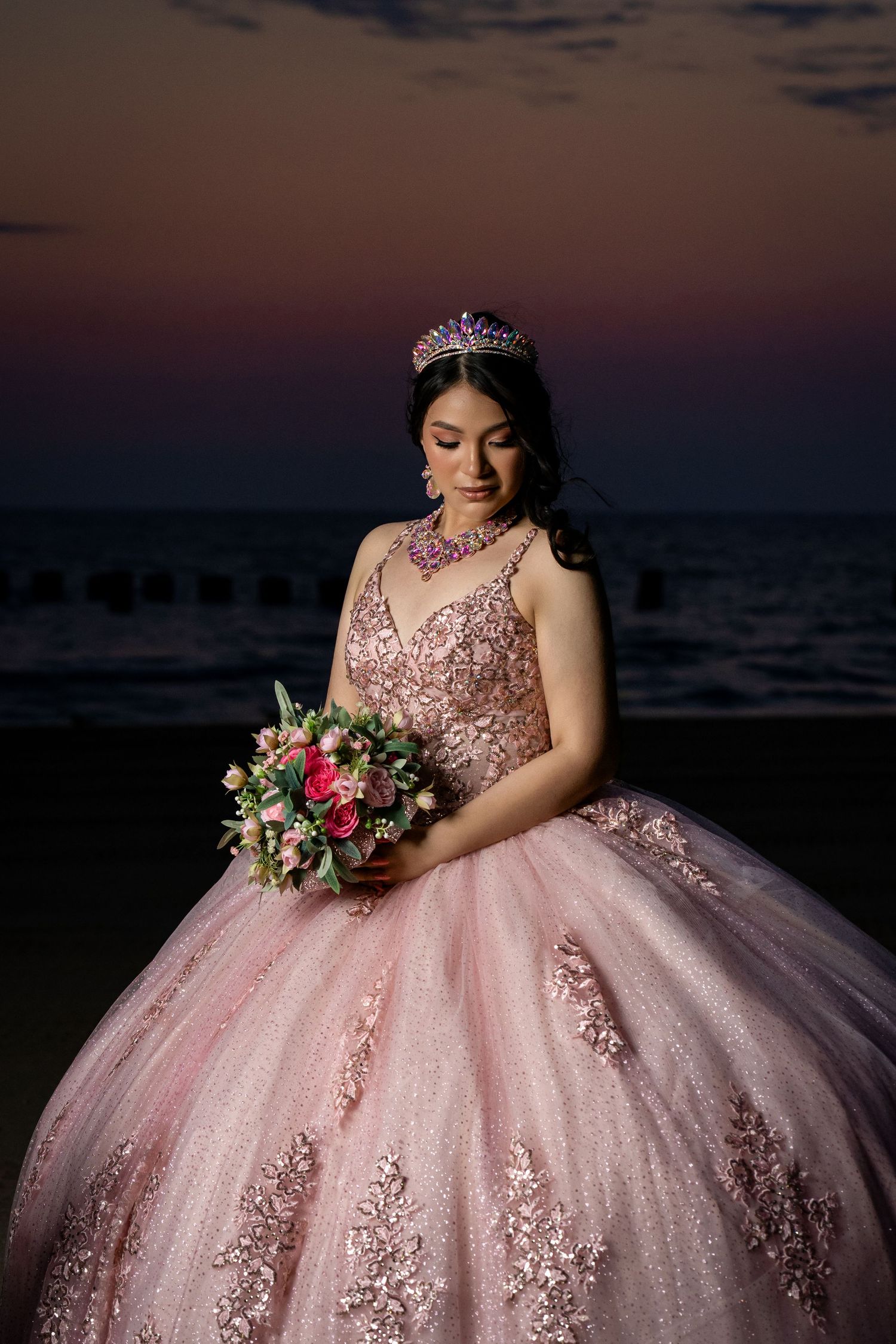A person in a pink ballgown with floral lace details poses against a sunset backdrop holding a colorful bouquet.