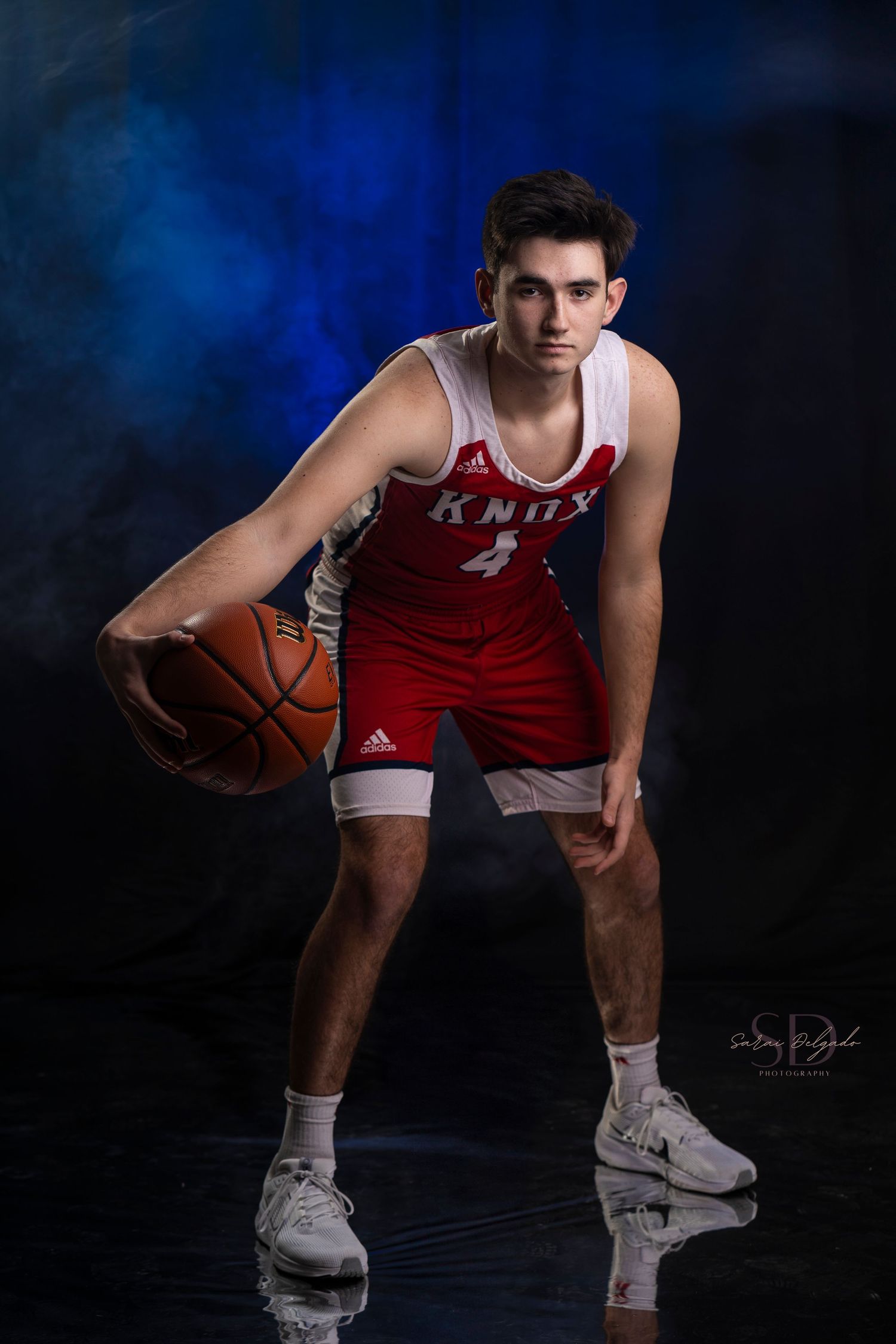 Basketball player in red and white RMU uniform poses in athletic stance with ball against dark blue backdrop.