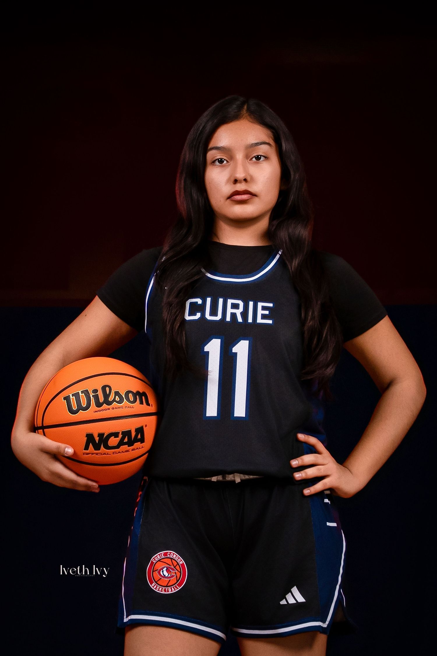 Basketball player poses in dark uniform holding orange ball against dramatic black background.