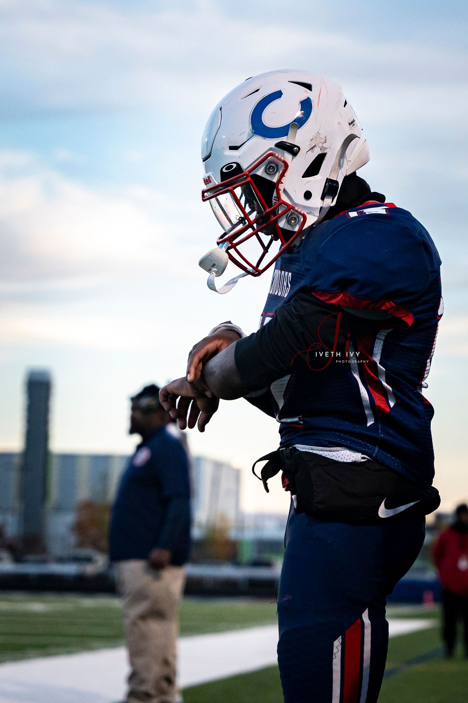 Football player in navy and red uniform holds ball while posing outdoors against city skyline and clouds.