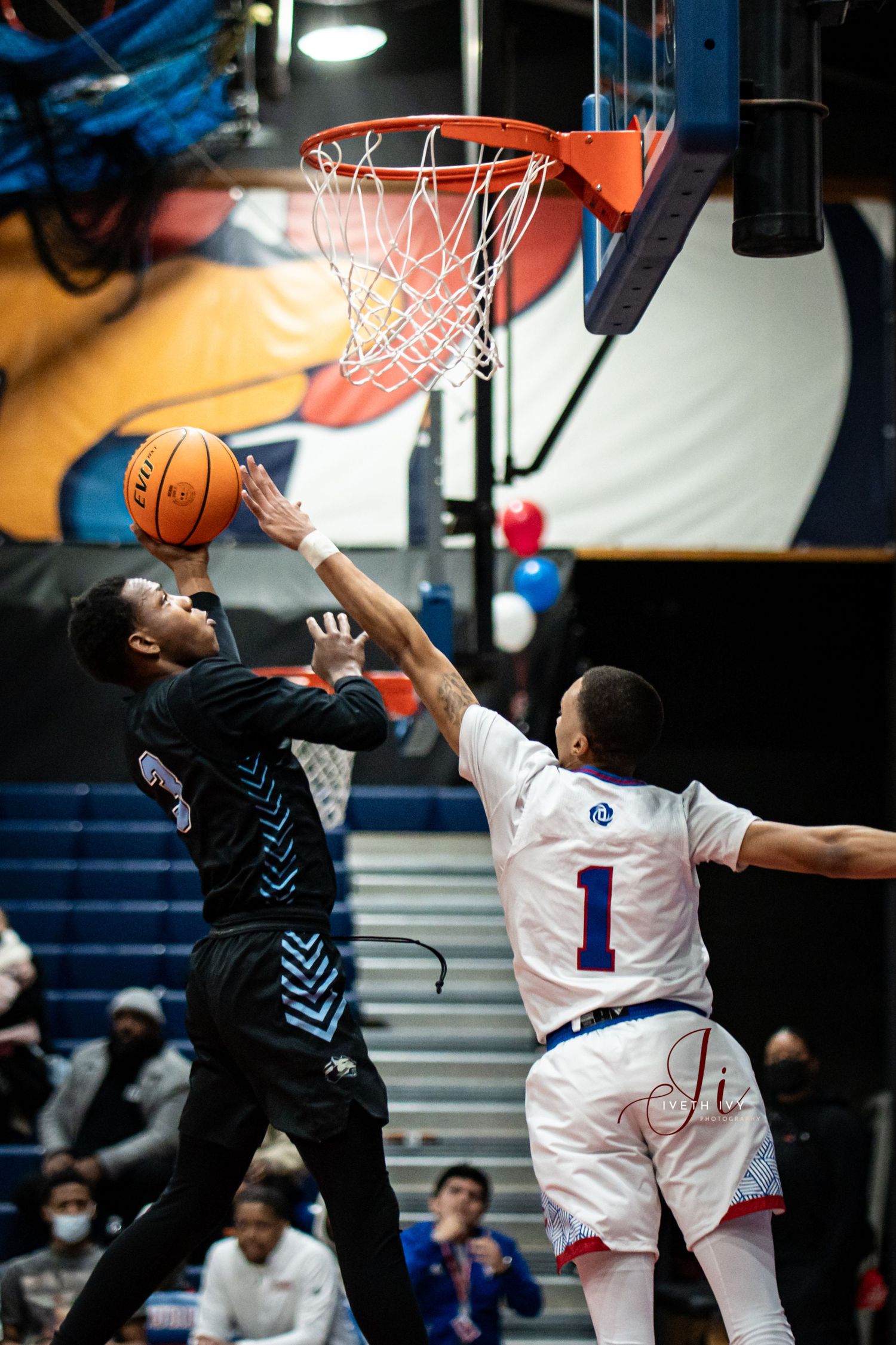 Basketball players leap for a rebound during an intense game with blue and white jerseys visible in the action sequence.