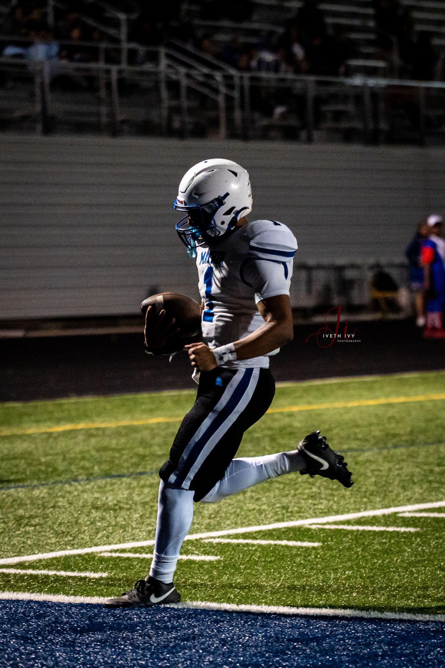Football player in white uniform leaping into blue end zone during night game.