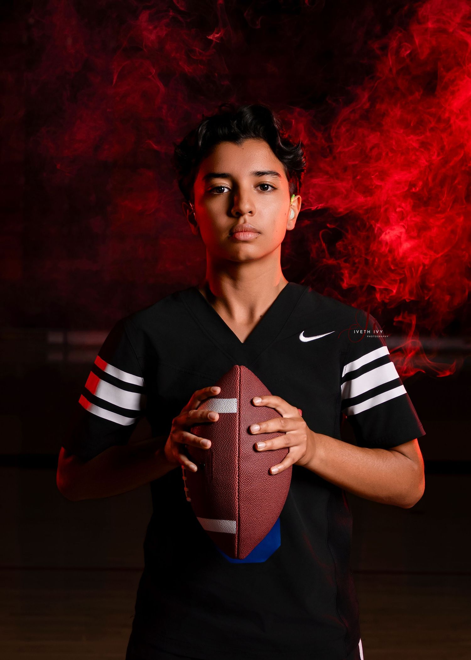 A young athlete in a black Nike jersey holds a football against a dramatic red smoke background.