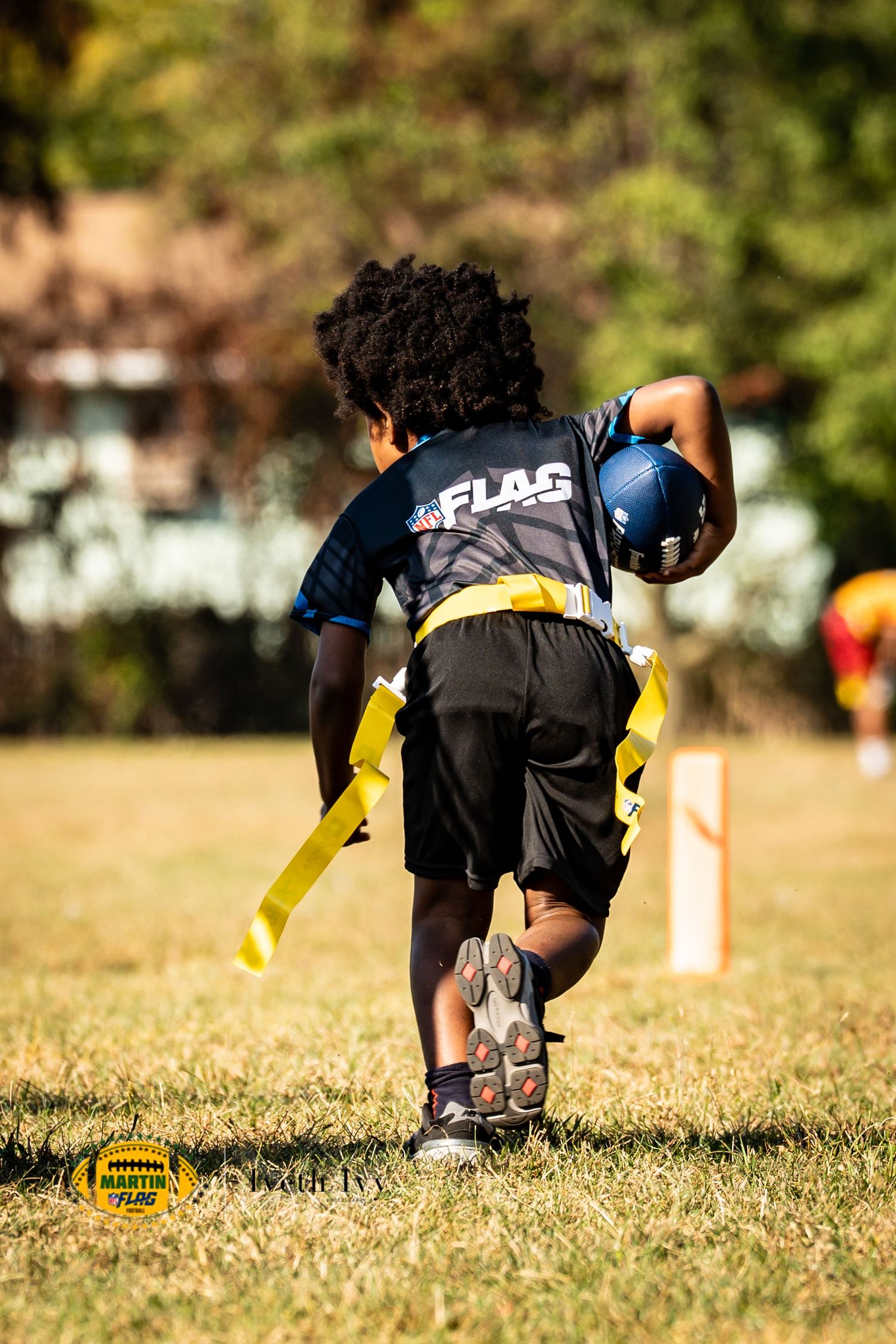 Youth flag football player in black and yellow uniform runs with a blue football during an outdoor game.