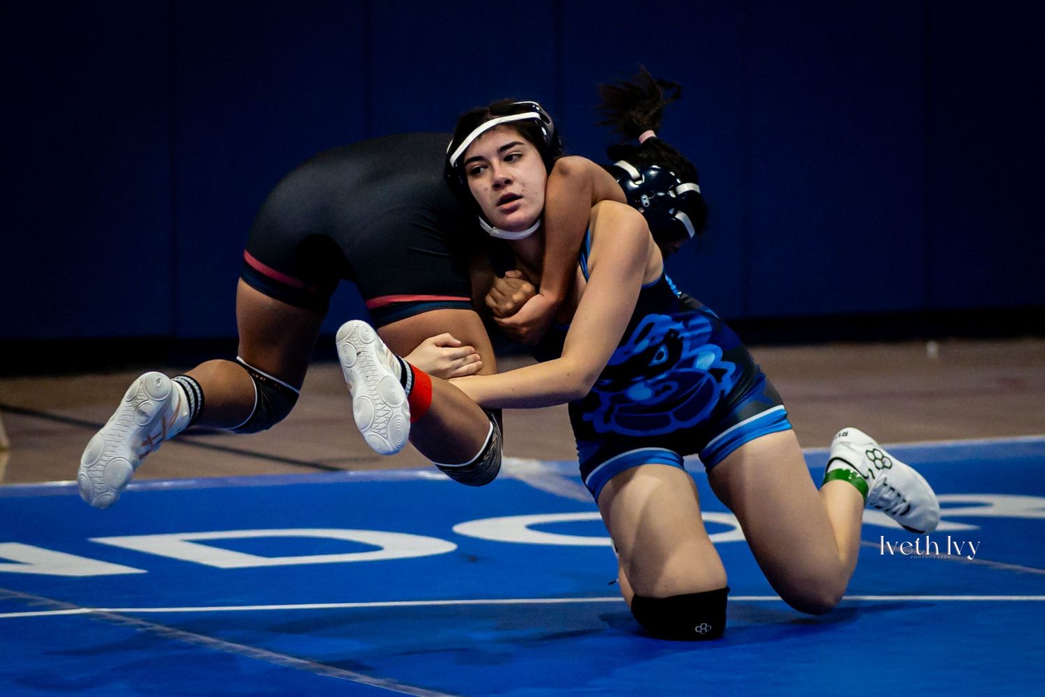 Two wrestlers compete on a blue mat during an intense grappling match at a wrestling tournament.