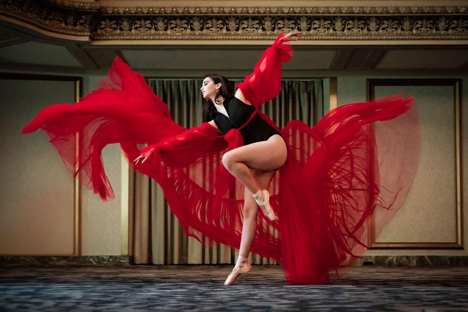 Dancer in black leotard leaps gracefully while flowing red fabric billows dramatically around them in an ornate hall.