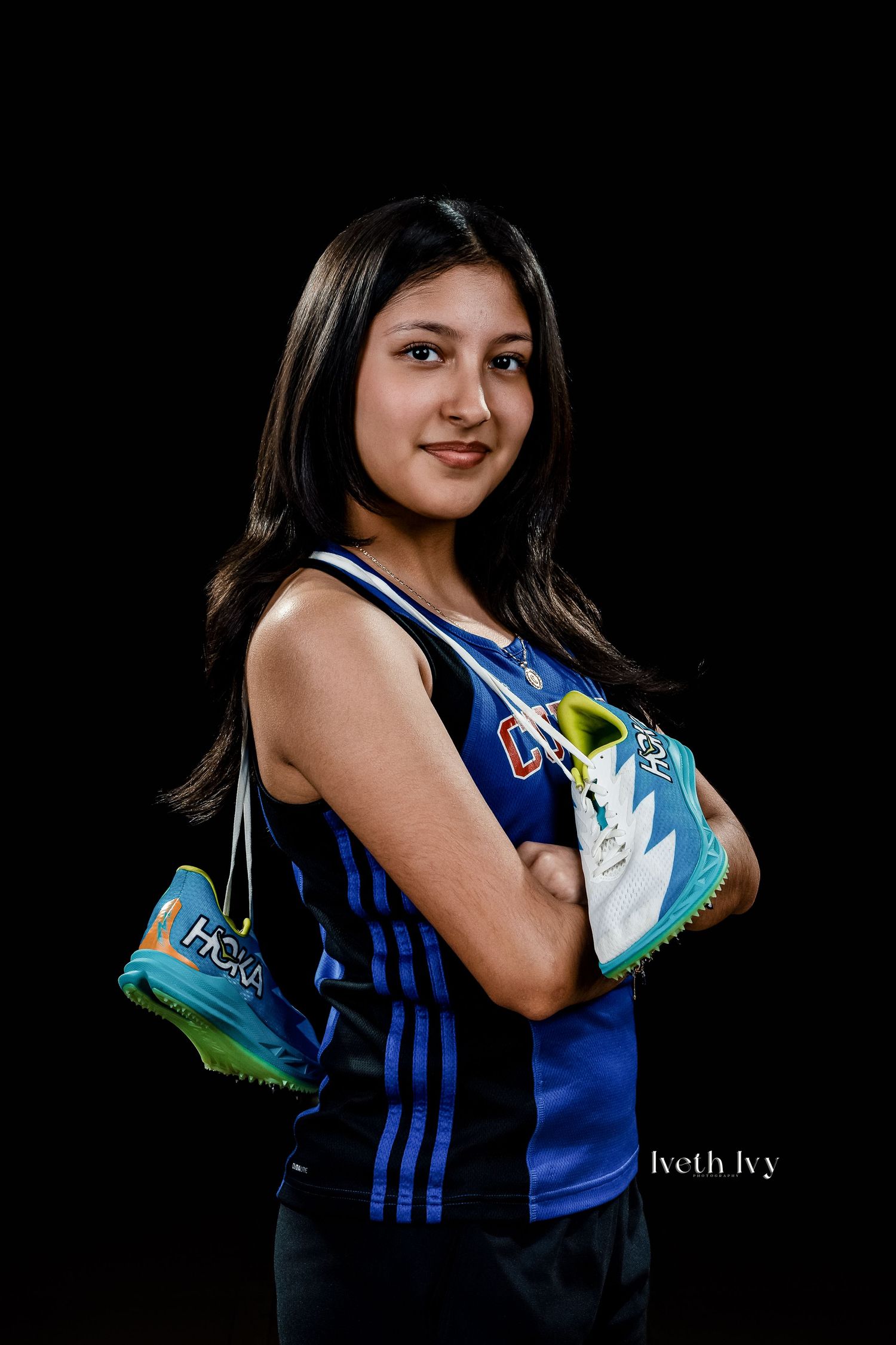 Young female athlete in blue sports jersey holds colorful track spikes with arms crossed against black background.