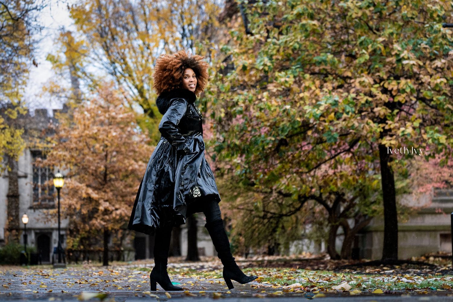 A person in a black coat and boots walks through a park with autumn leaves falling from trees in the background.