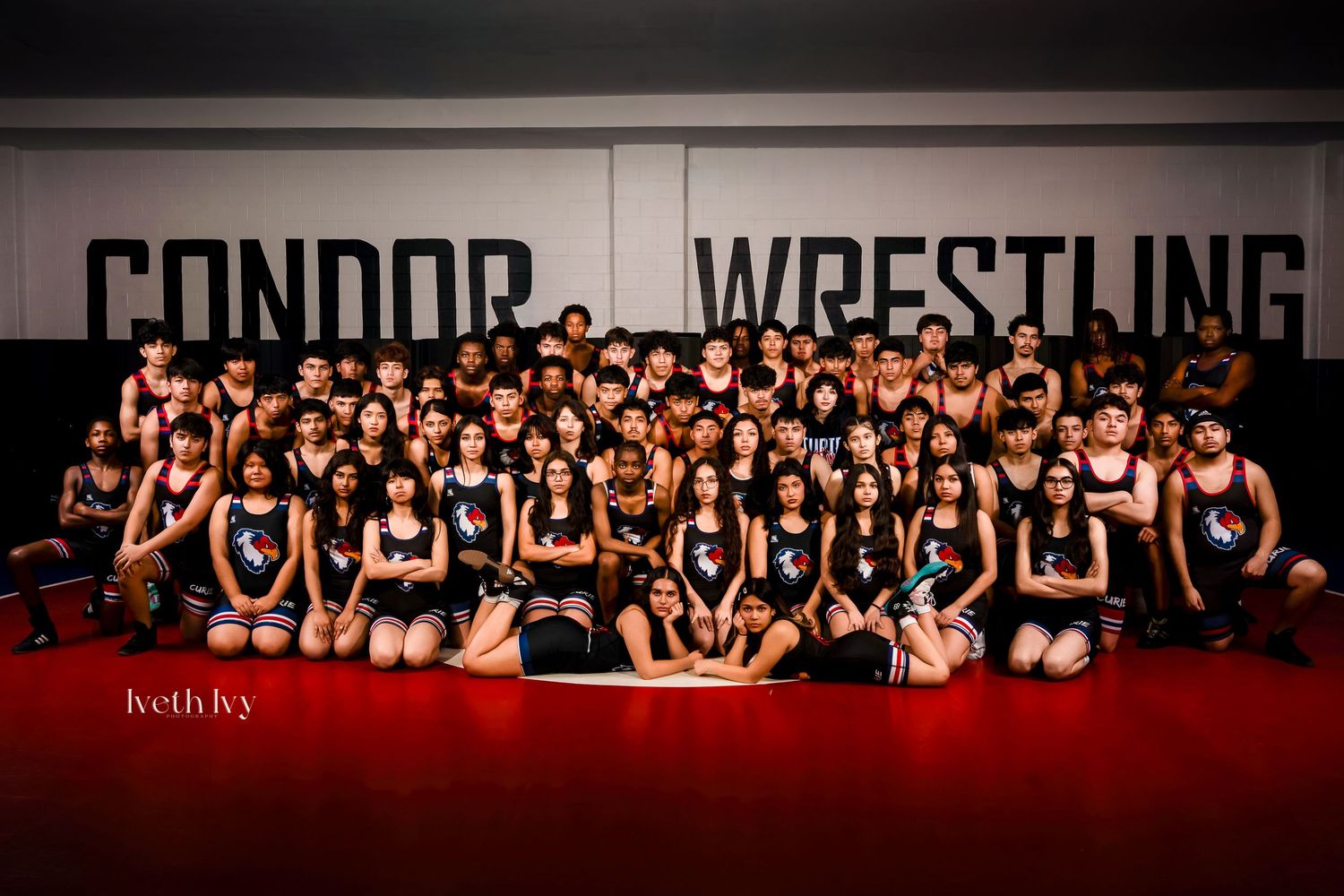 Large group of wrestlers in black uniforms pose together on red mat in front of Condor Wrestling wall sign.