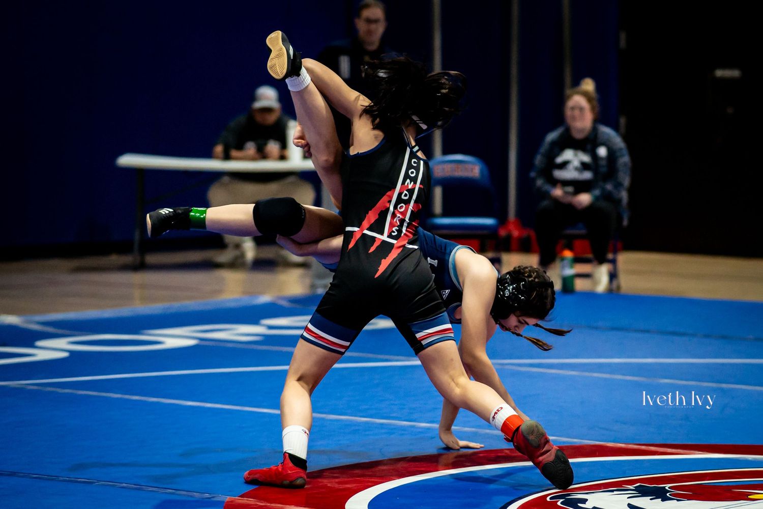 Wrestlers compete on blue and red wrestling mats during a competitive match.