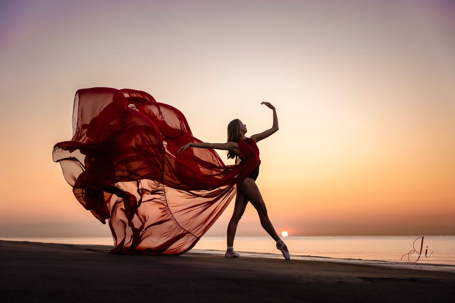 Silhouette of dancer with flowing red dress against sunset on beach.