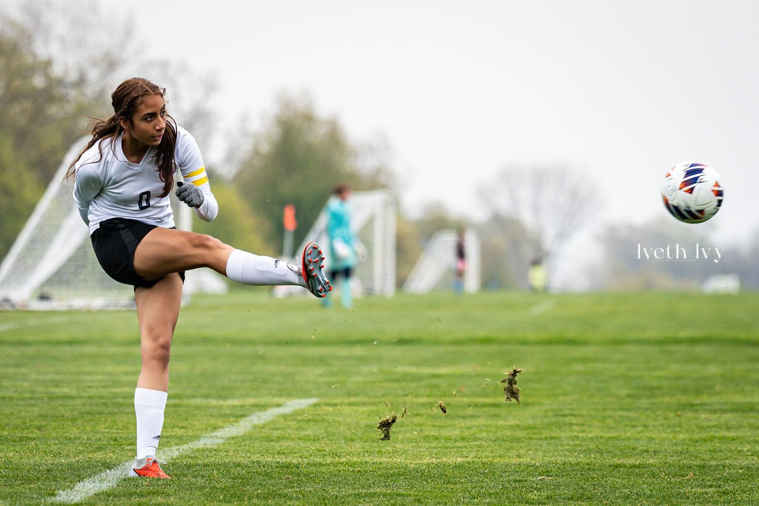 Female soccer player kicking ball mid-air on green field with goal in background during outdoor match.