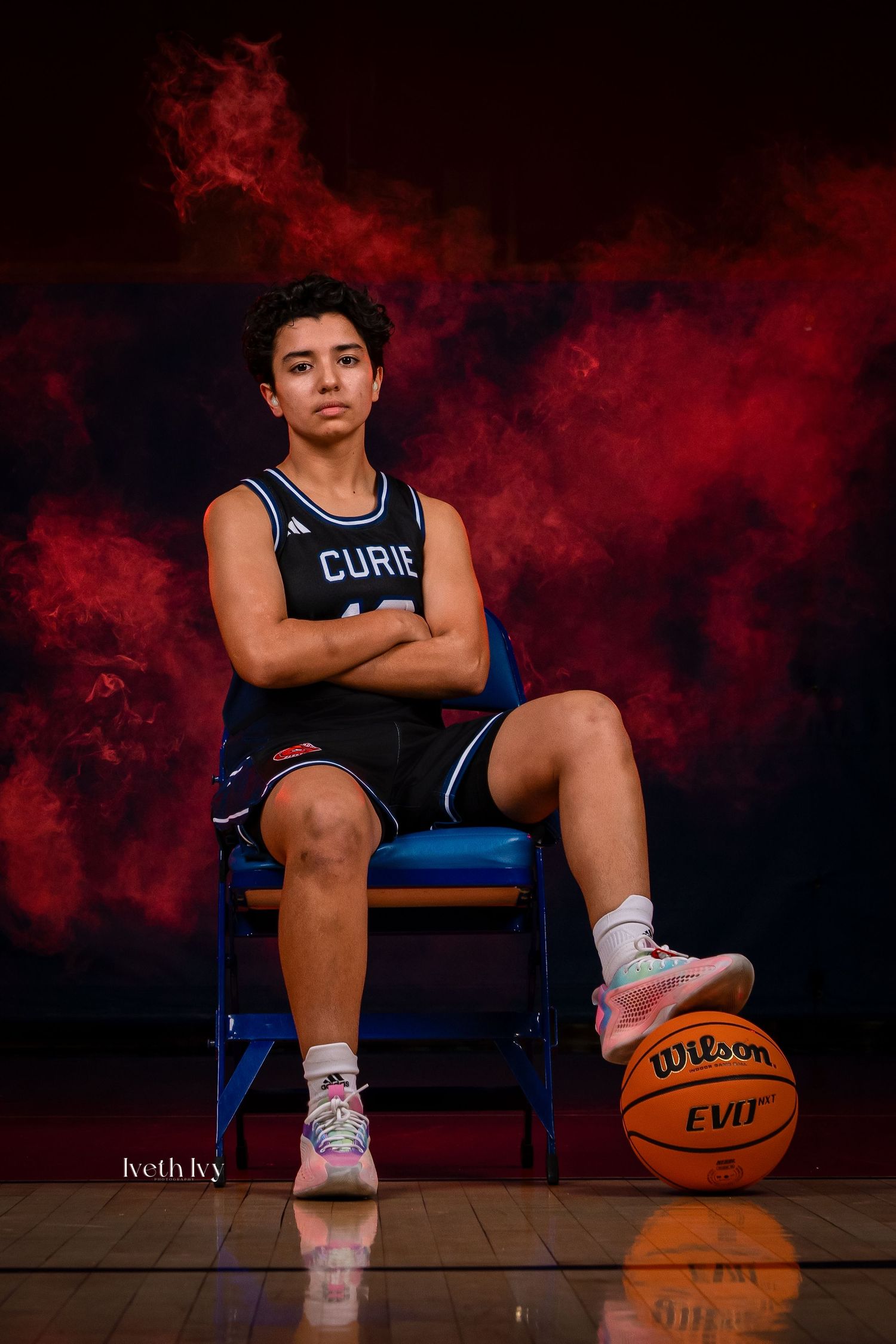 Basketball player sits on blue chair with Wilson basketball against dramatic red smoke backdrop for sports portrait.