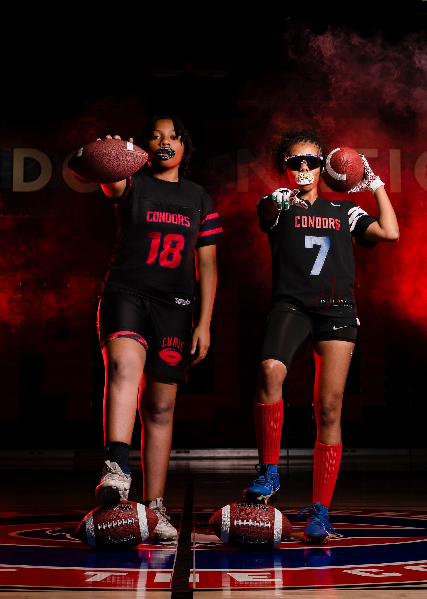 Athletic legs and feet in sports gear stand on footballs against a dramatic red and black backdrop during a photoshoot.