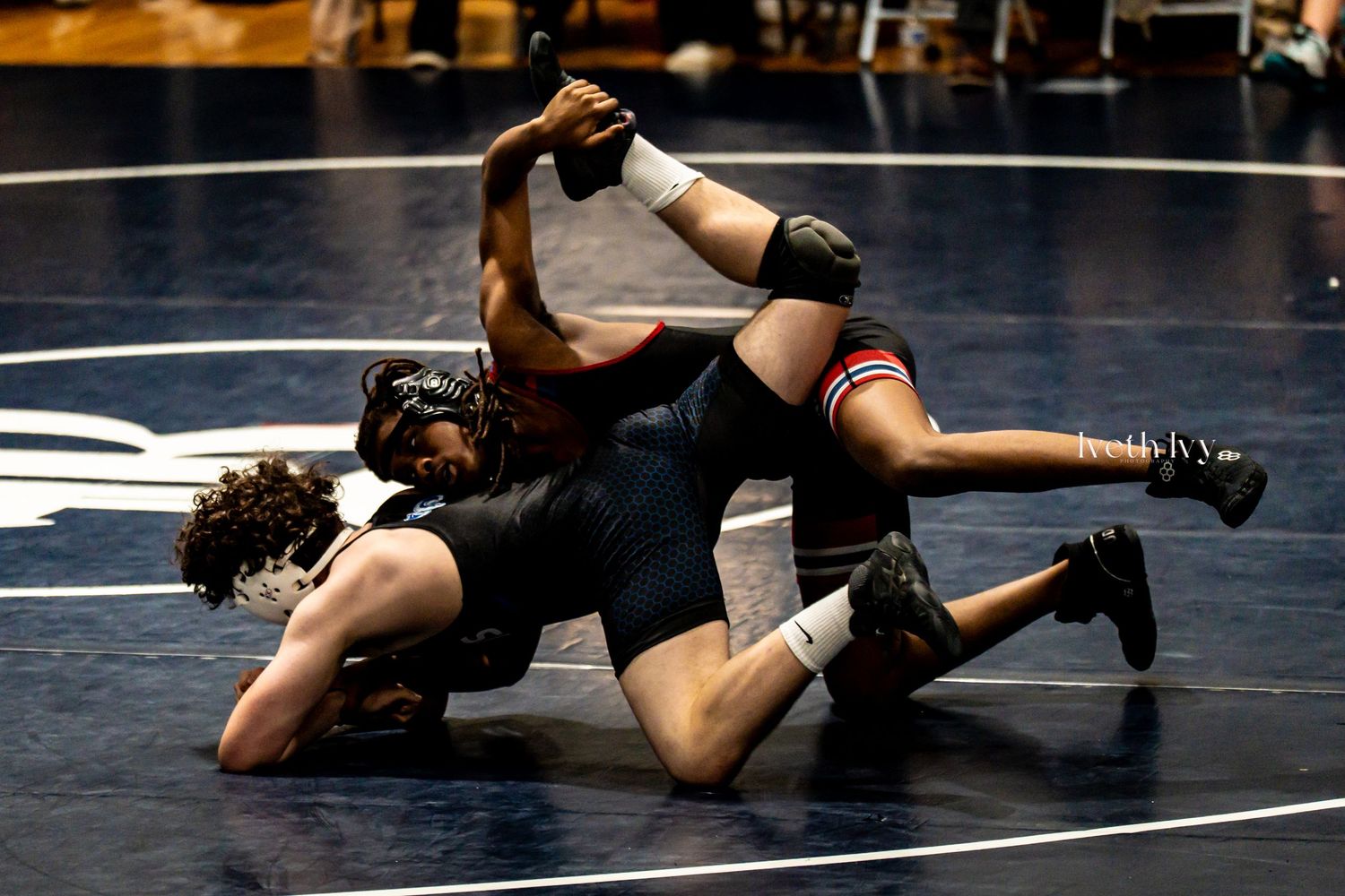 Two wrestlers compete in an intense match on a dark blue wrestling mat during a competitive bout.