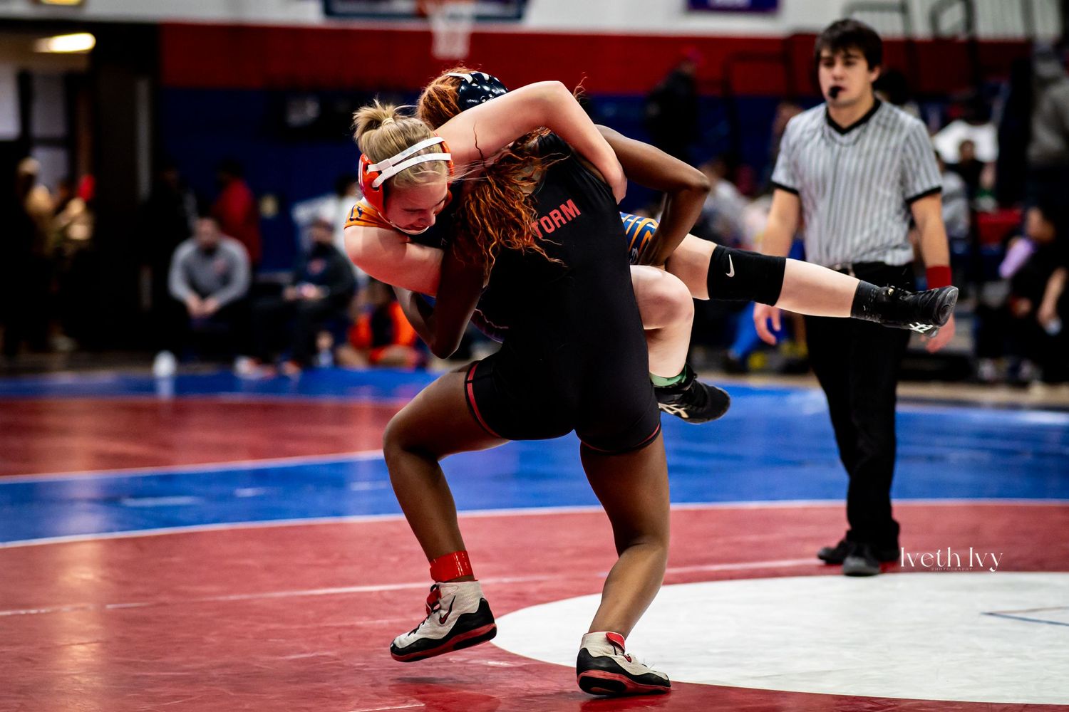 Two wrestlers grapple intensely on a red and blue competition mat during a wrestling match.