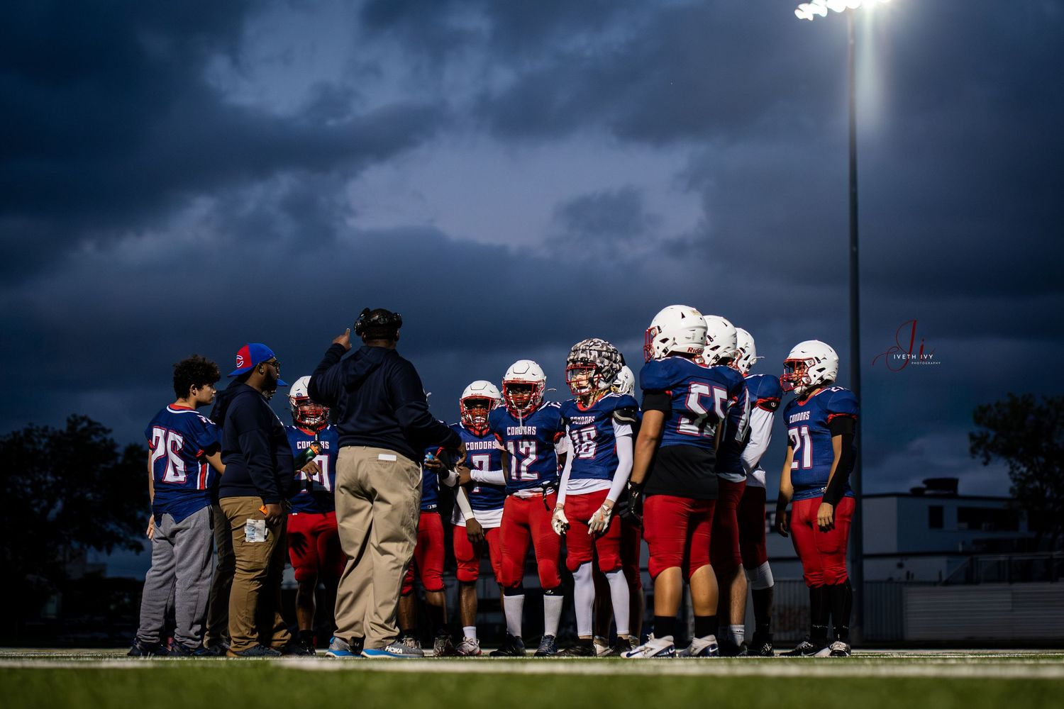 Football players and coaches gather under stormy skies during an evening game as stadium lights illuminate the field.