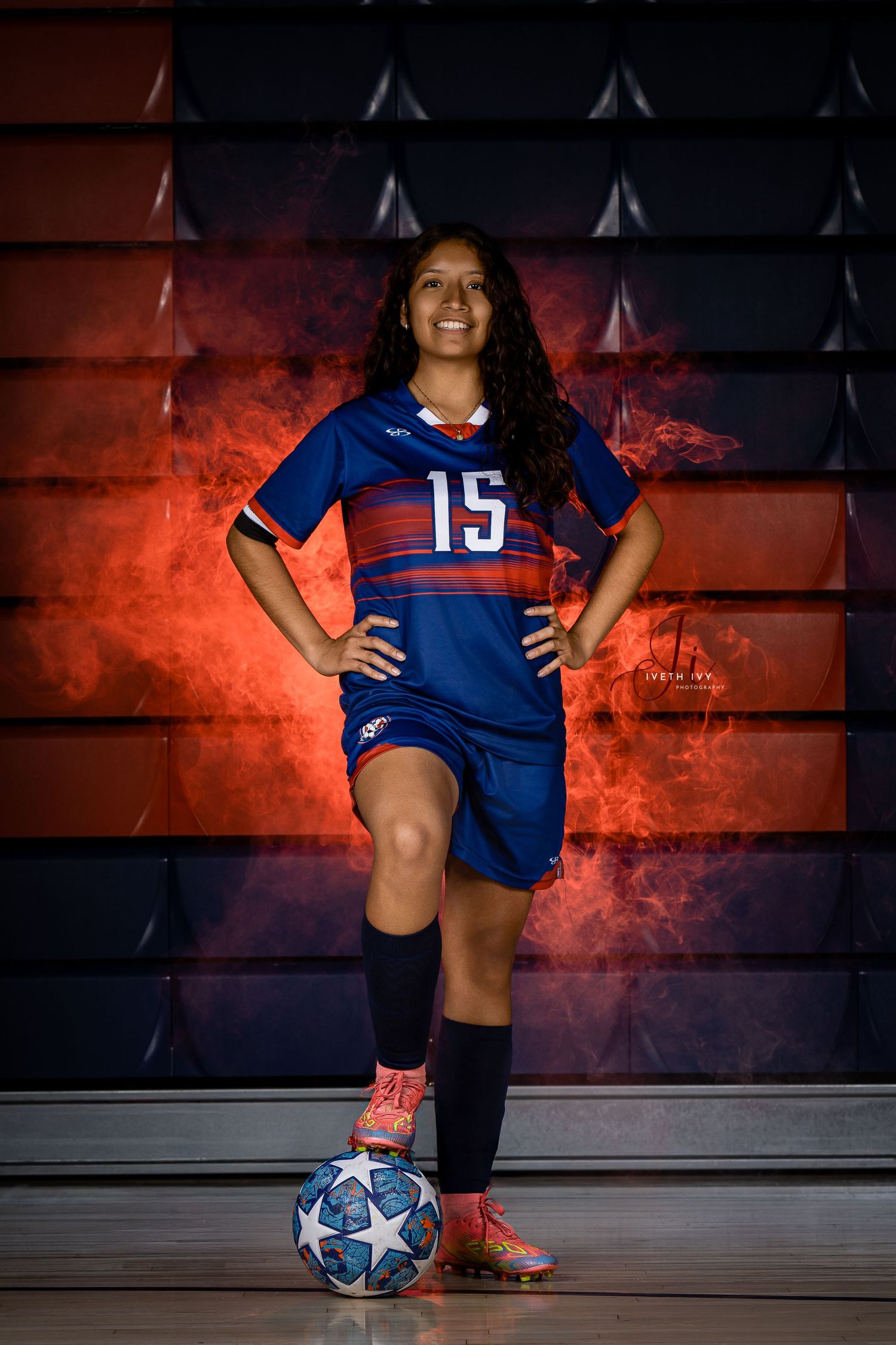 Soccer athlete poses with ball in blue and red uniform against dramatic red-lit background and American flag display.