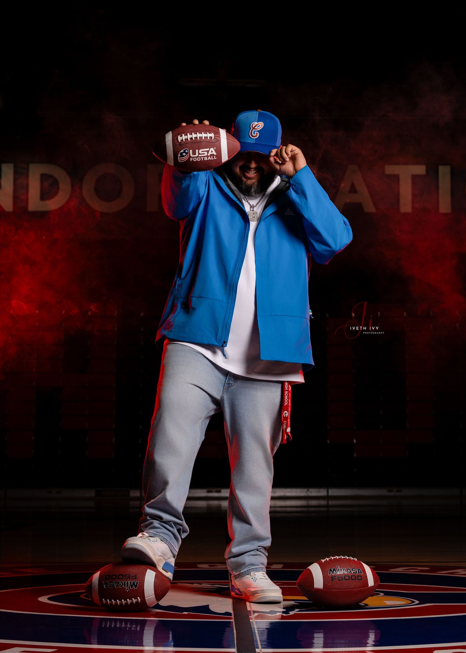 A person wearing blue jacket and gray sweatpants poses with footballs in a dramatic red-lit studio setting.