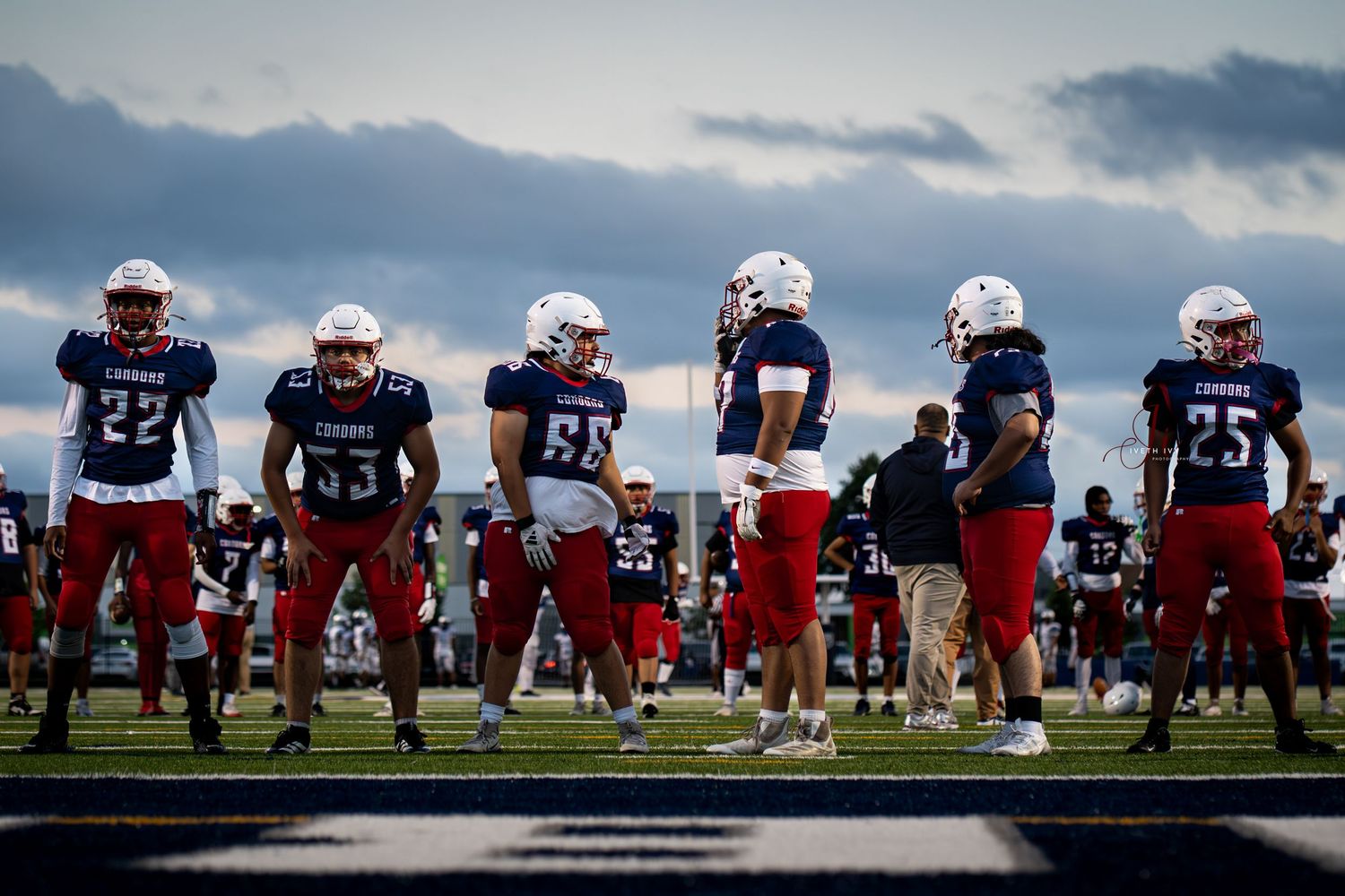 Football players line up in formation during practice on a sunny day with dramatic clouds overhead.