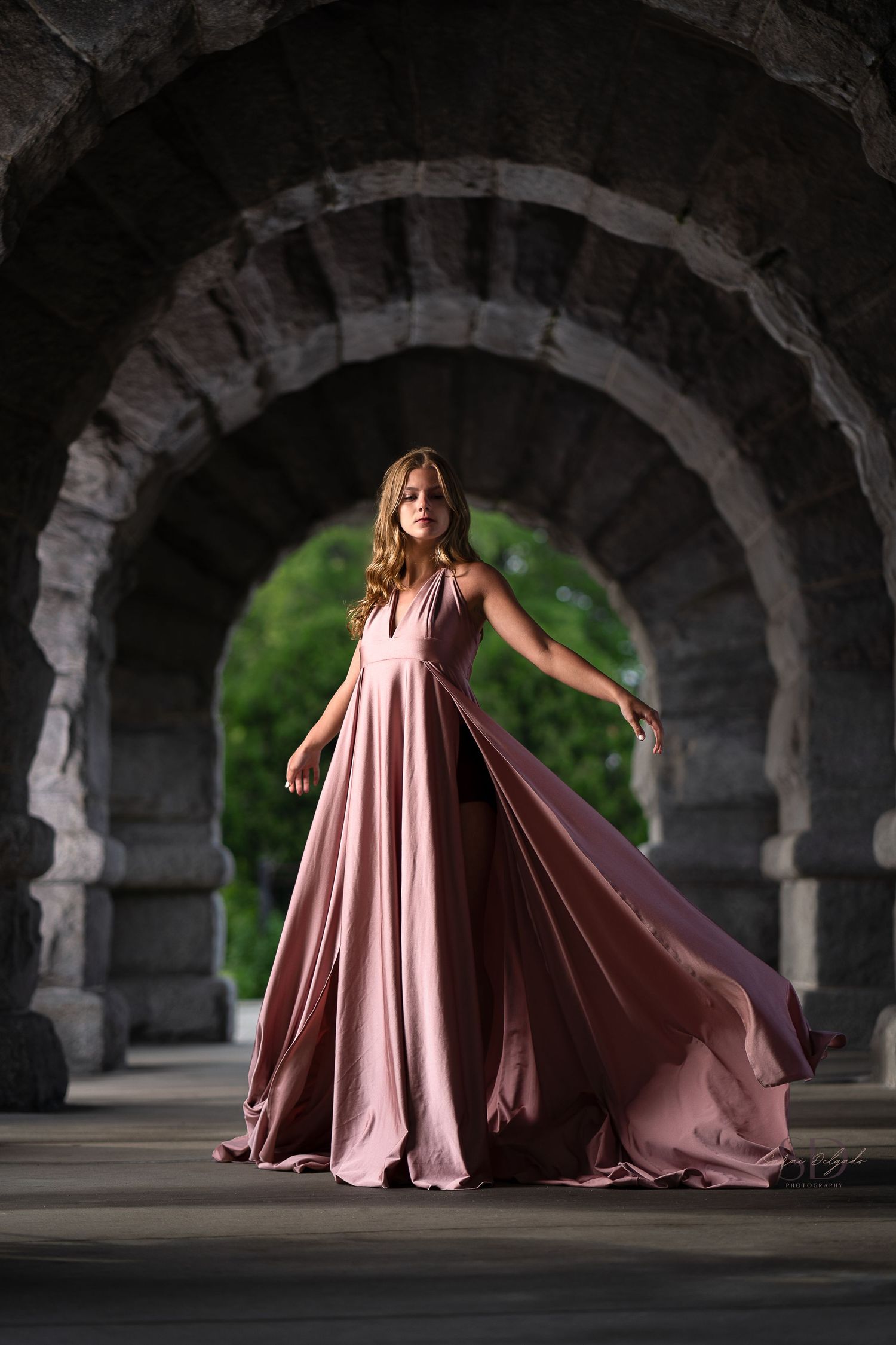 Flowy mauve evening gown flowing dramatically under stone archway in artistic fashion photography.