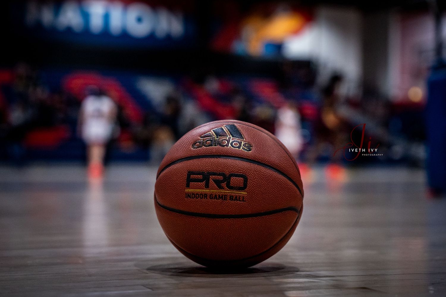A close-up of a basketball on a glossy court floor with blurred stadium lights and crowd in the background.