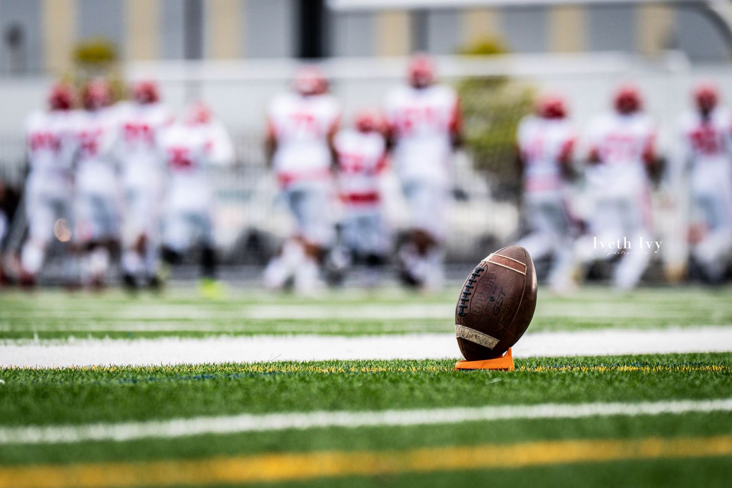 A football lies on the turf of a stadium field with players and crowd visible in the background.