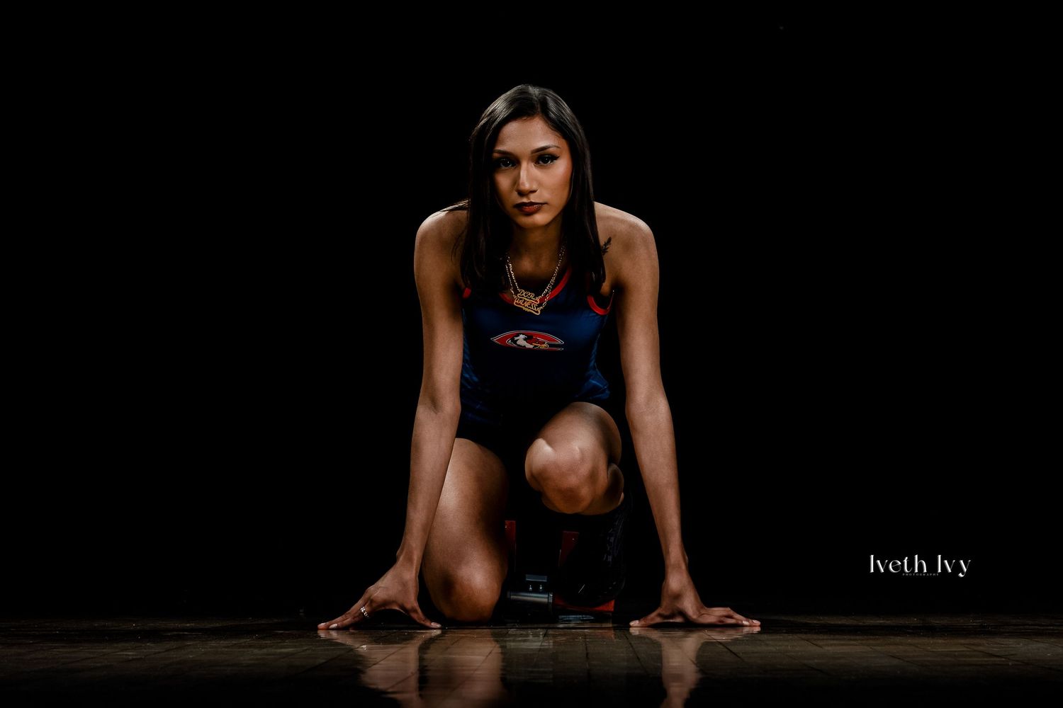 Female athlete in starting position on a dark studio floor wearing a blue athletic outfit.
