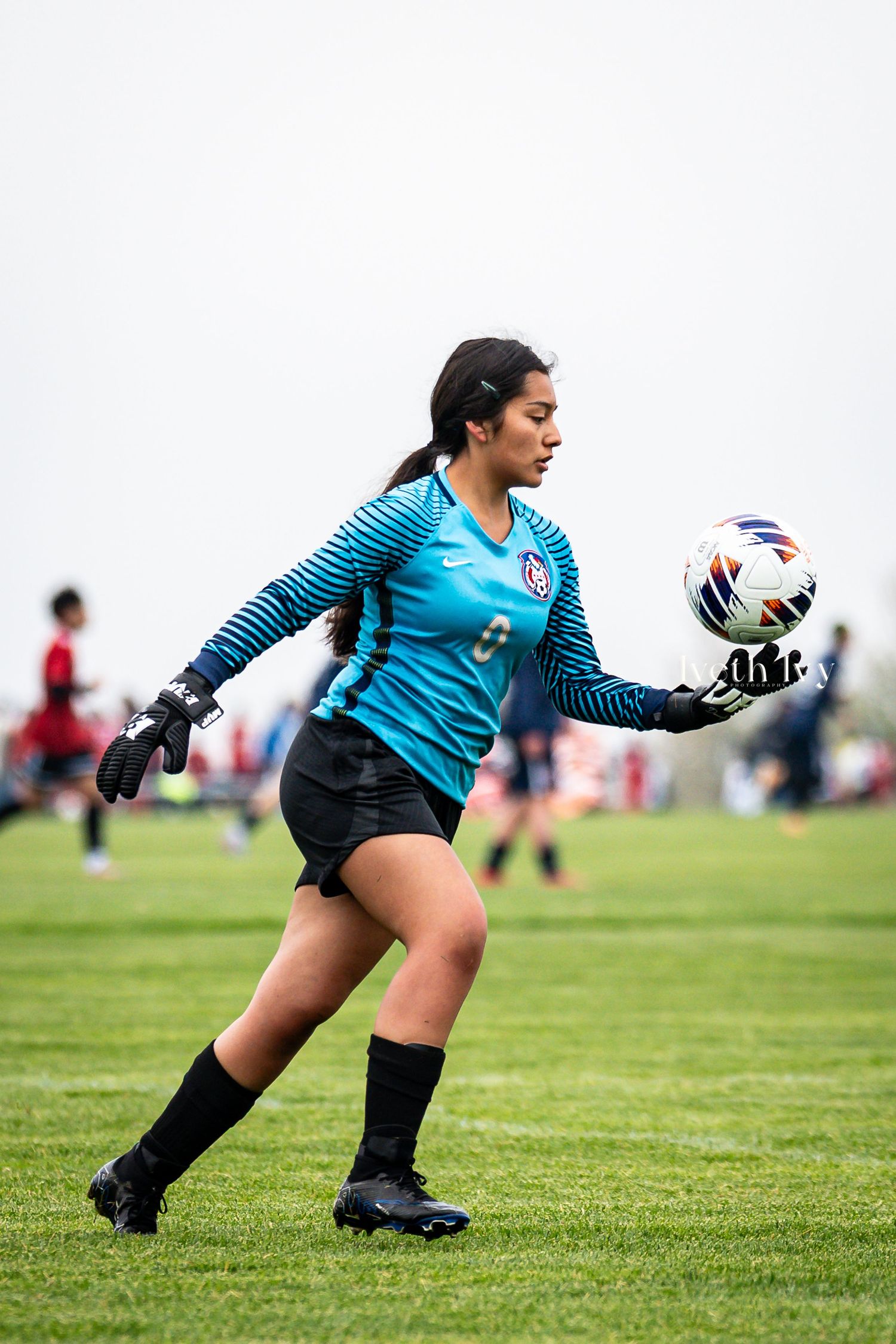 Female goalkeeper in teal Nike jersey holding a soccer ball on a grass field during a game.