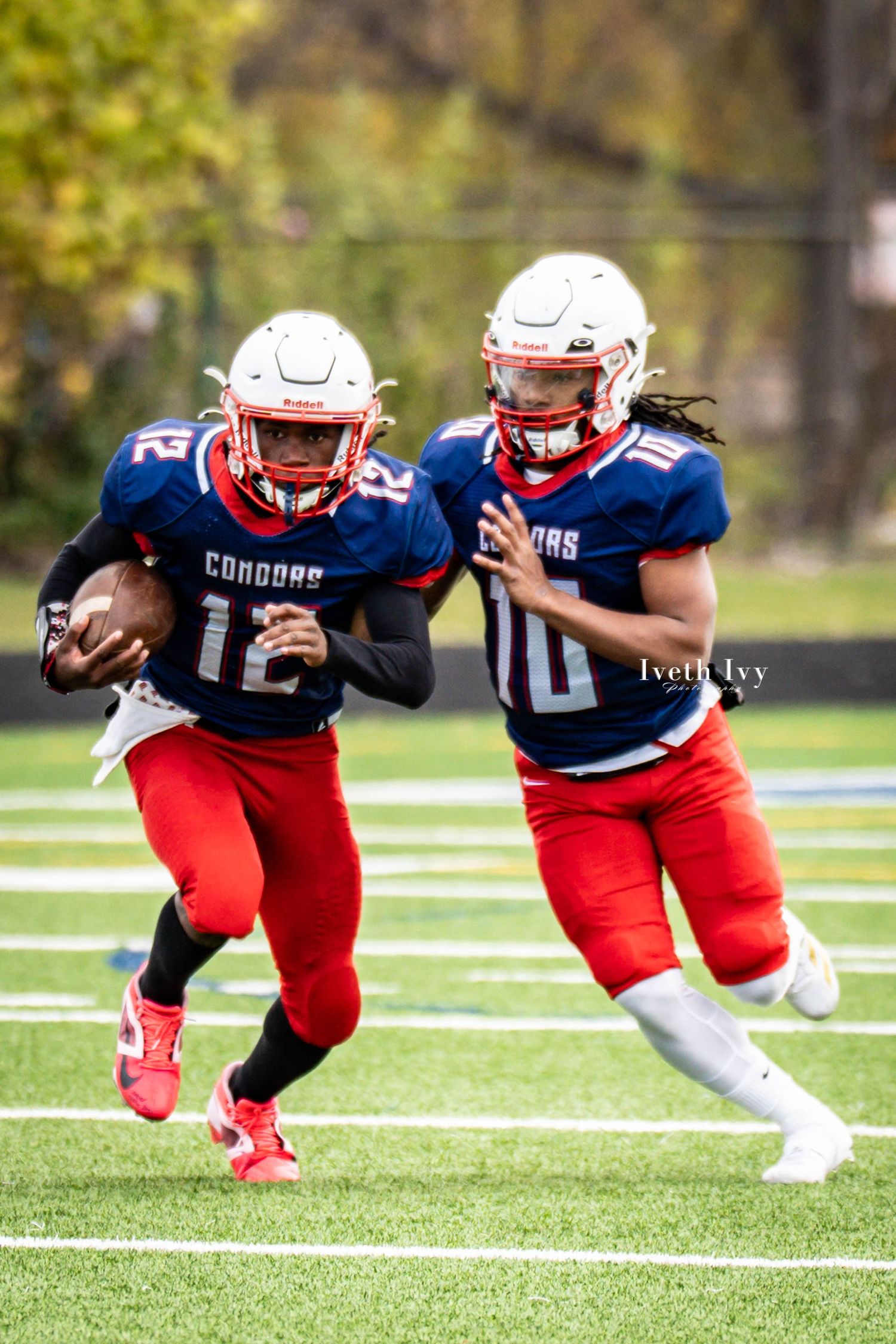 Two football players in red and blue uniforms practicing blocking drills on grass field.