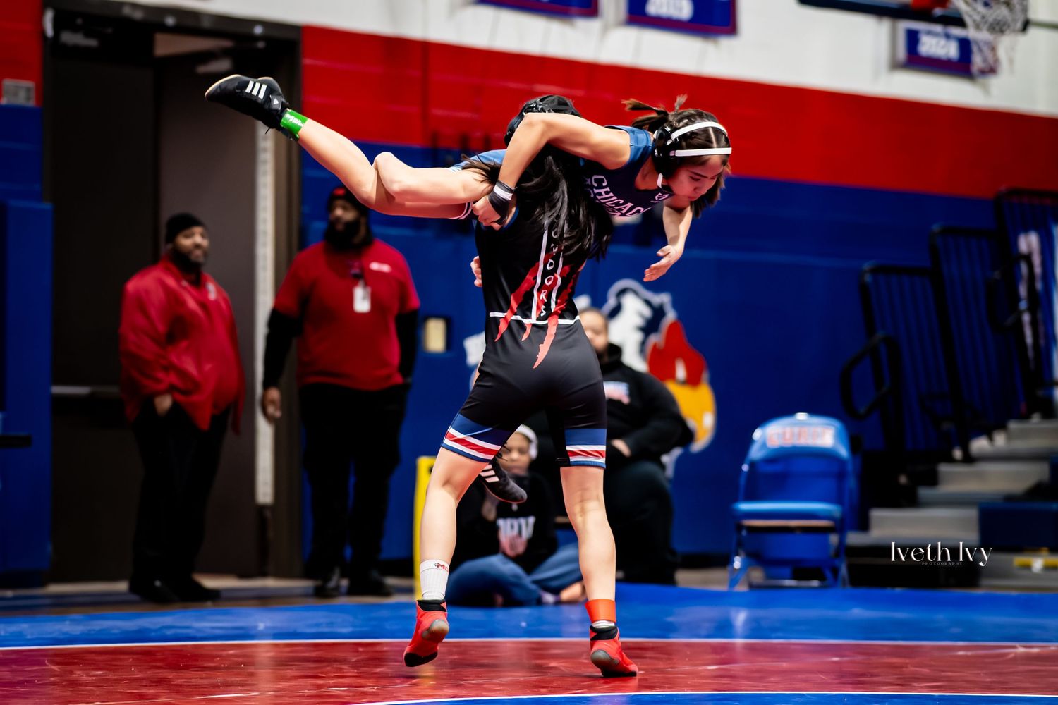 Two wrestlers compete in a high school match with one athlete lifting and controlling the other on a blue mat.