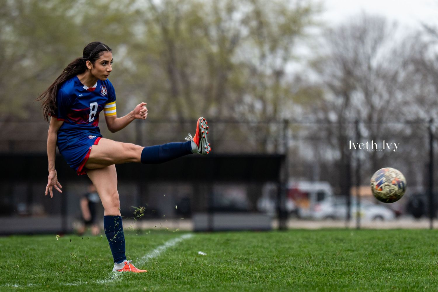Female soccer player in blue and red uniform kicking ball on green field.